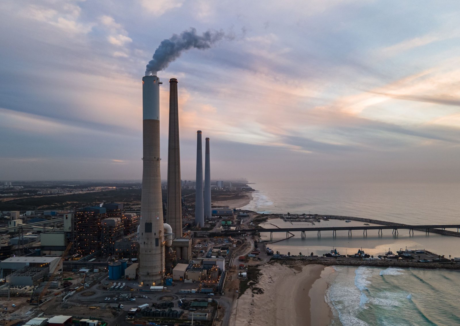 Aerial view of an industrial plant by the ocean with smoke stacks at sunset, capturing an industrial and serene contrast.